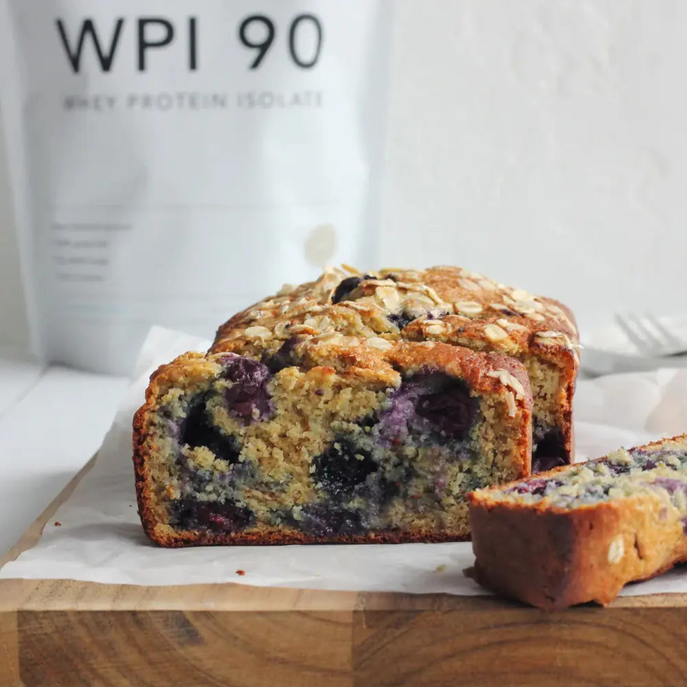 Blueberry protein loaf cake with oats on top on a wooden cutting board, sliced piece in foreground and white WPI 90 whey protein isolate bag blurred in background.
