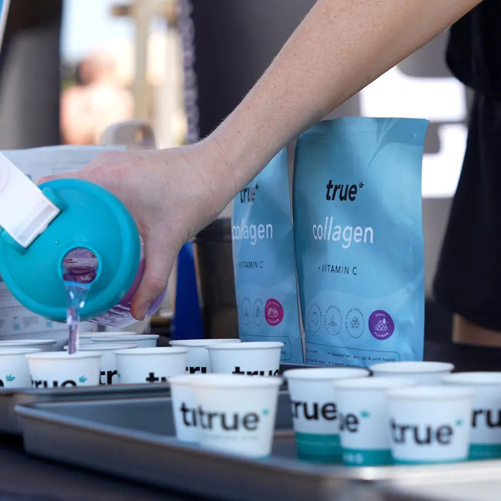Person pouring a purple drink into small branded sample cups at an outdoor market stall with two blue "true collagen + Vitamin C" product pouches visible in the background.