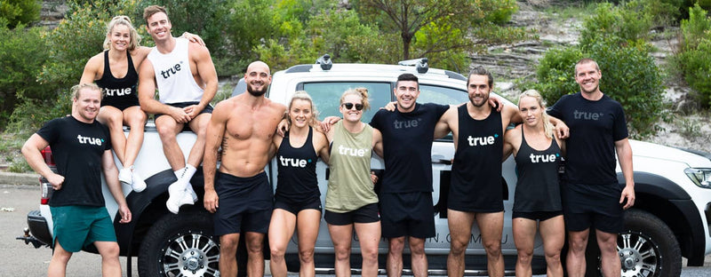 Group photo of ten fit True-branded trainers and athletes posing together in front of a white 4x4 truck outdoors in Australia, wearing True logo fitness apparel for a team/bootcamp marketing image