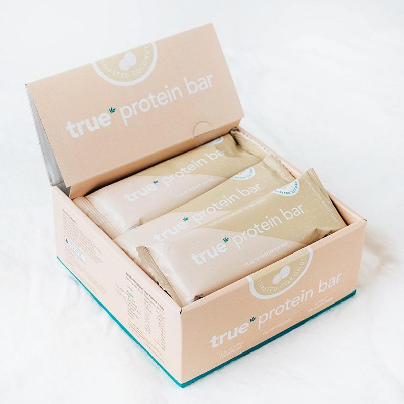 Open box of True Protein Bar toasted coconut flavor with beige individually wrapped protein bars on a white background