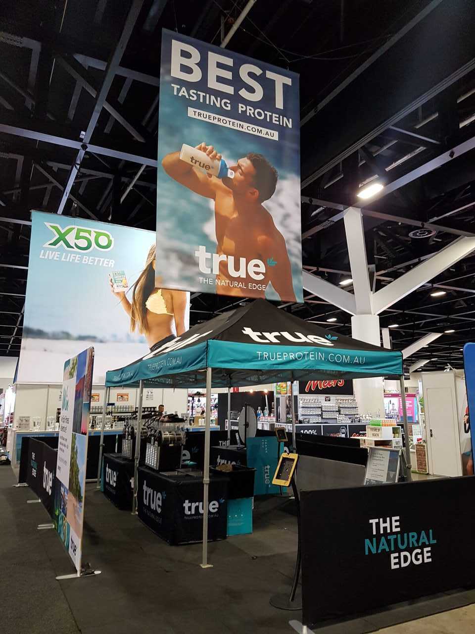 True Protein convention booth at an Australian health expo — turquoise canopy with "trueprotein.com.au", large hanging "BEST TASTING PROTEIN" banner, "The Natural Edge" signage and product sampling counters.