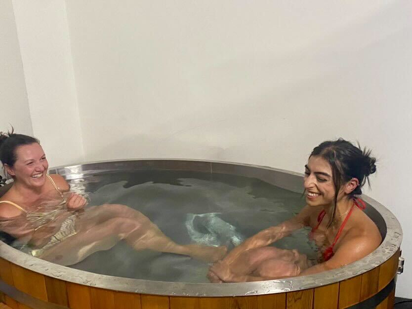 Two women laughing in swimsuits while sitting in an indoor wooden ice bath / cold plunge tub for wellness and recovery.