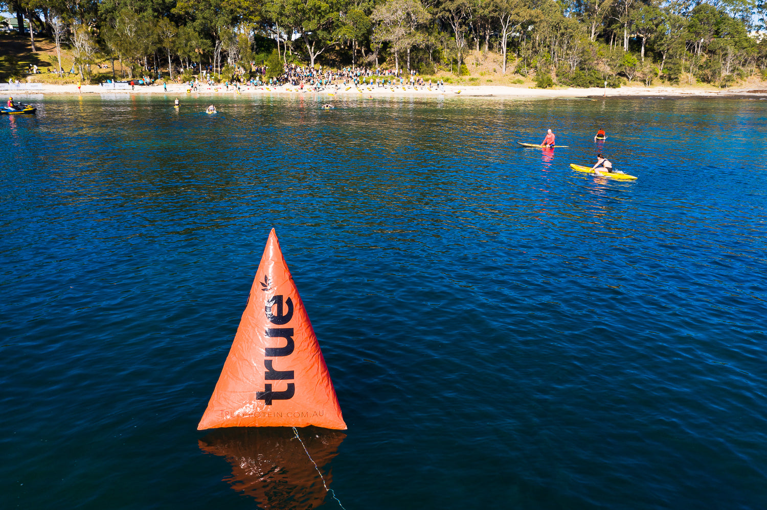 Orange triangular race buoy branded "True" floating in blue bay water with kayakers paddling nearby and a sandy shoreline crowded with spectators and eucalyptus trees during The Bay Games in Australia.