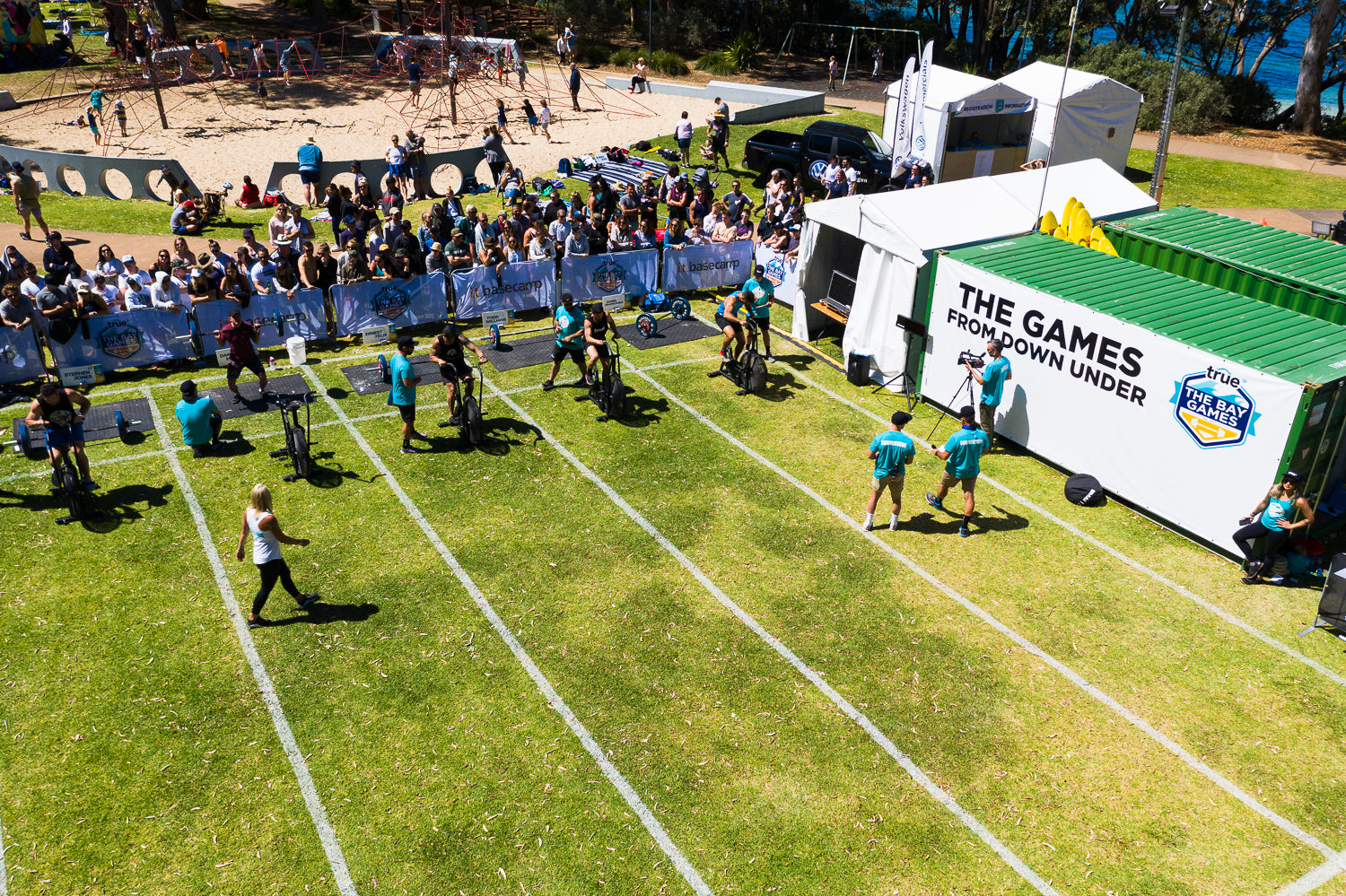 Aerial view of The Bay Games outdoor fitness competition on a grassy coastal park in Australia, showing athletes on exercise bikes and rowing stations racing in marked lanes, event staff, spectators behind barriers, sponsor tents and a white container branded "The Games From Down Under."