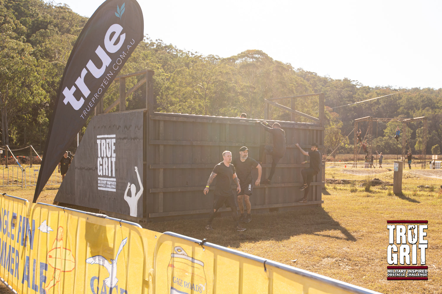Participants climbing a wooden wall obstacle at a True Grit military-inspired obstacle course event in rural Australia, black True Protein flag and logo visible, sunlit bushland background.