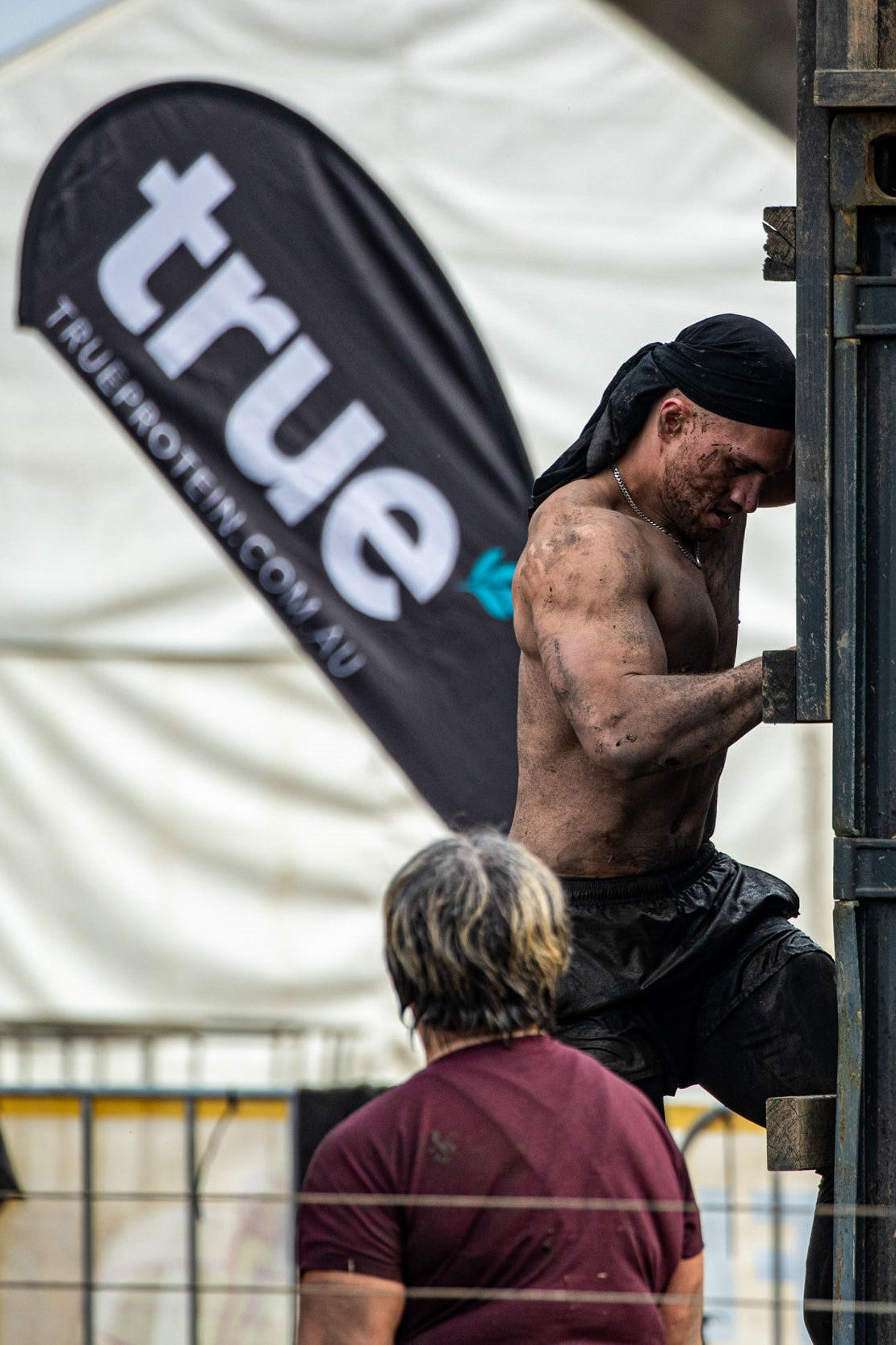 Muddy male obstacle-course racer climbing a wooden wall at a True Grit outdoor fitness event, branded "True Protein" flag visible — obstacle race in Australia.