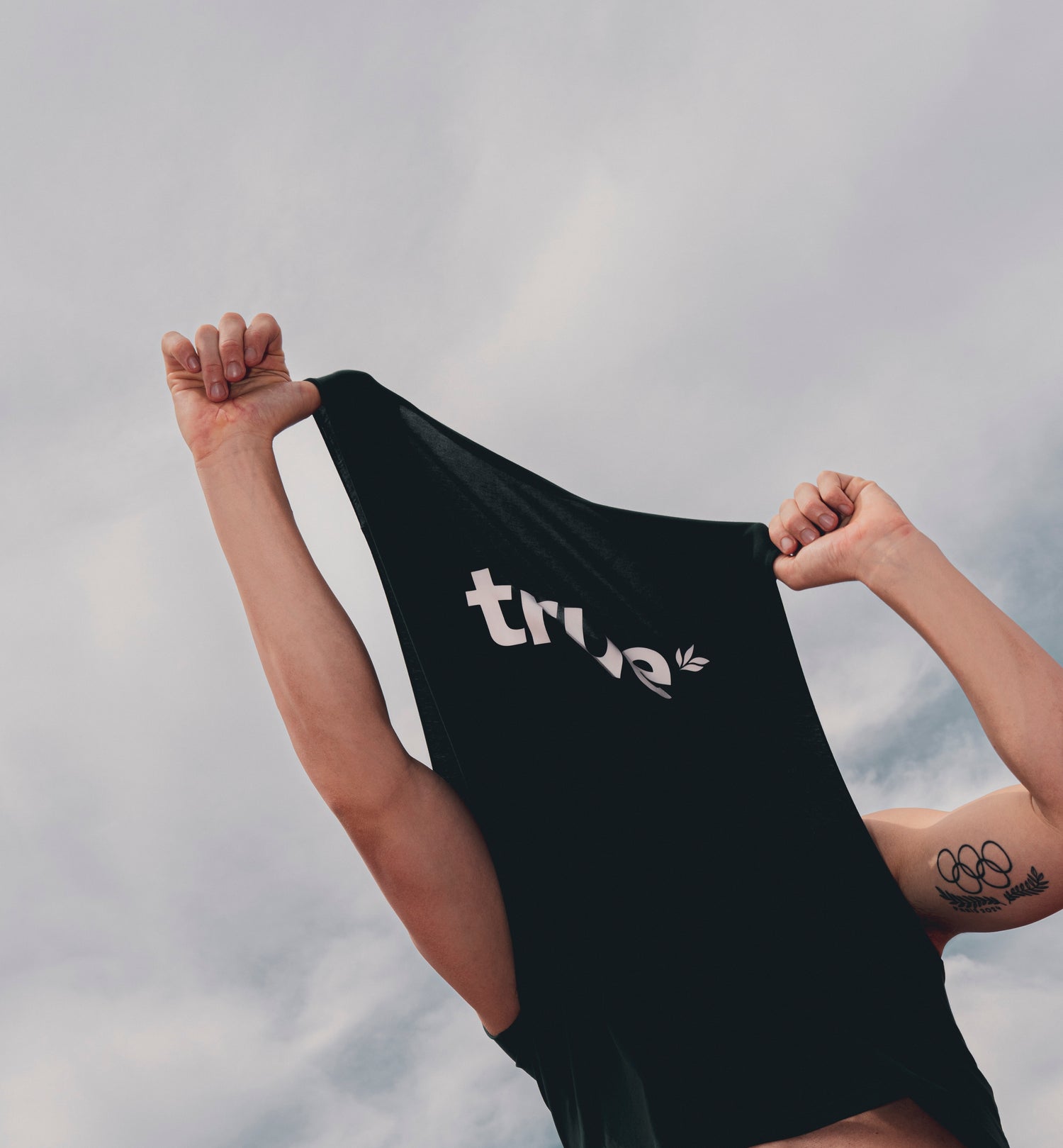 Black True Protein t-shirt with white "true" logo being pulled over a fit model's head, raised muscular arms and Olympic-rings tattoo visible against a cloudy outdoor sky — fitness apparel product shot.