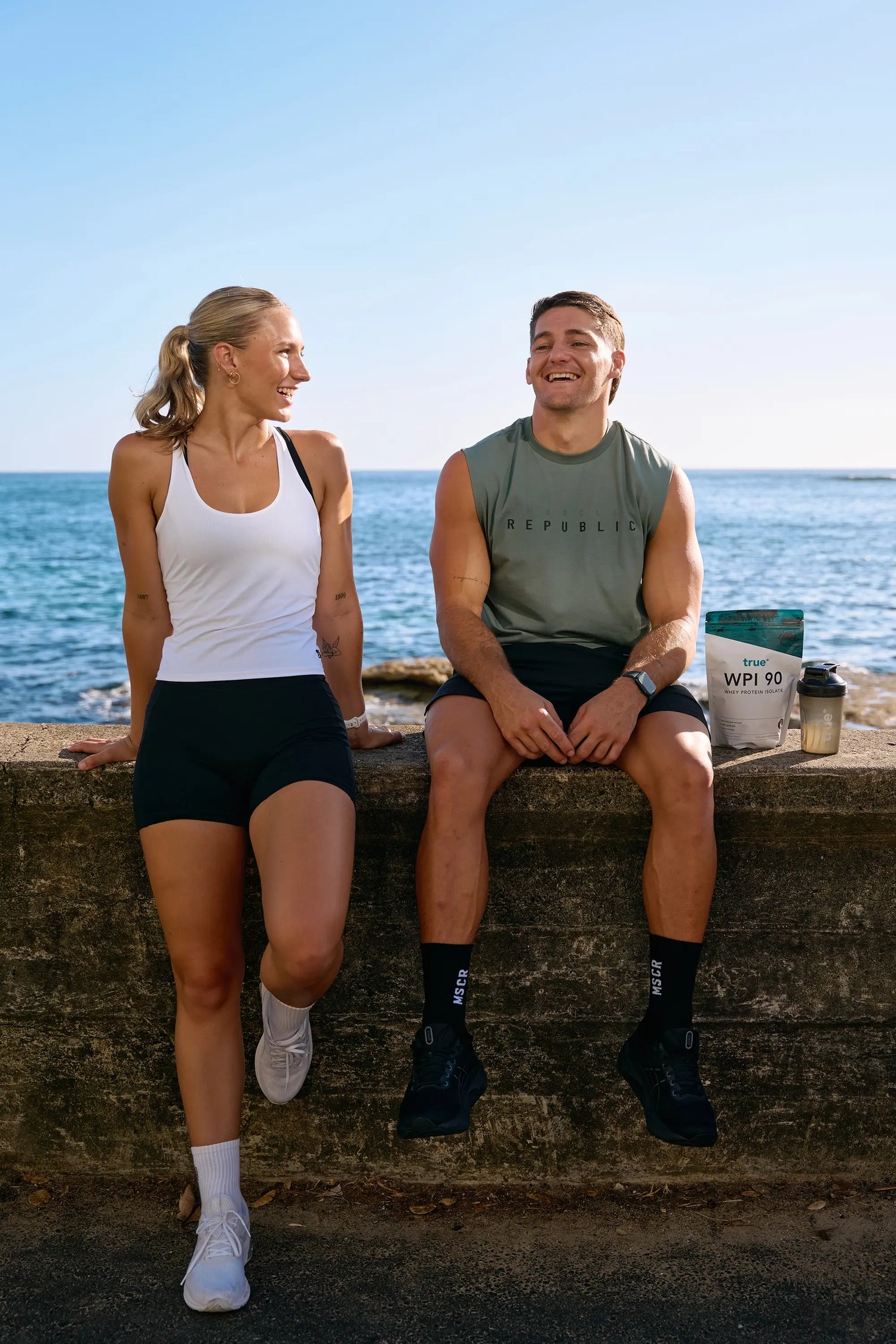 Two fit adults in workout clothing sitting on an oceanfront seawall, laughing beside a True WPI 90 whey protein bag and shaker—outdoor fitness, beachside workout lifestyle image.