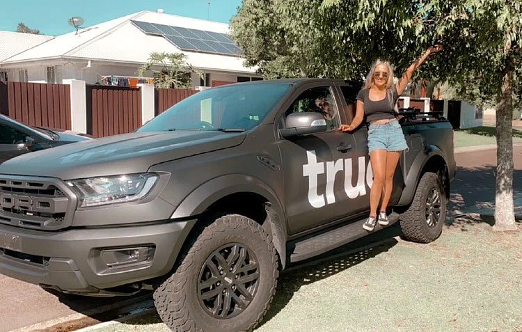 Matte black Ford Ranger Raptor pickup parked on a suburban street with solar-panel roof houses; a smiling woman in sunglasses, black tank top and denim shorts stands on the truck’s side step beside large off-road tires and a visible "tru" decal.