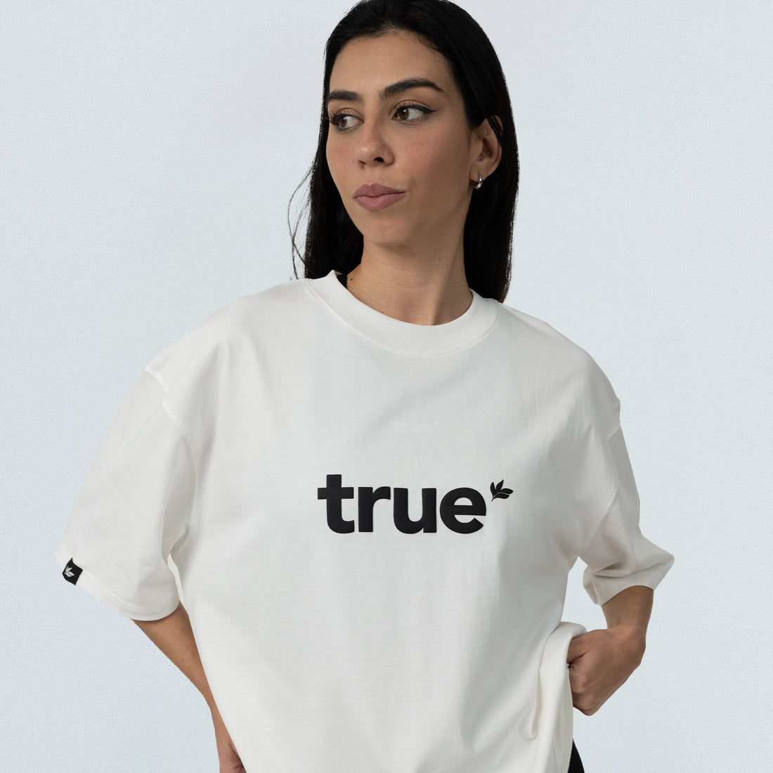 Female model wearing white unisex oversized cotton t-shirt with black "true" logo, front product shot on light gray background.
