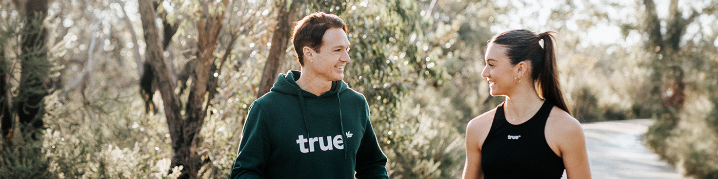 Two people wearing True-branded activewear walking and smiling on a sunlit wooded trail, outdoor fitness lifestyle, nature walk.
