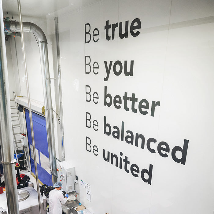 Cleanroom production facility interior with worker in white coat at machinery and a large wall mural reading “Be true, Be you, Be better, Be balanced, Be united” — food/manufacturing plant production line.