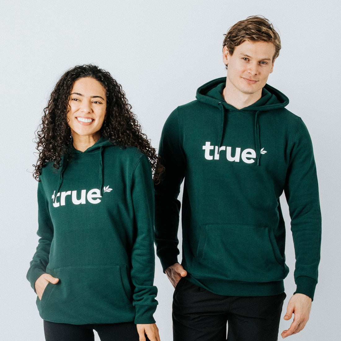 Two models wearing forest-green unisex organic cotton hoodies with white "true" logo and front kangaroo pocket — casual eco-friendly sweatshirt for men and women.