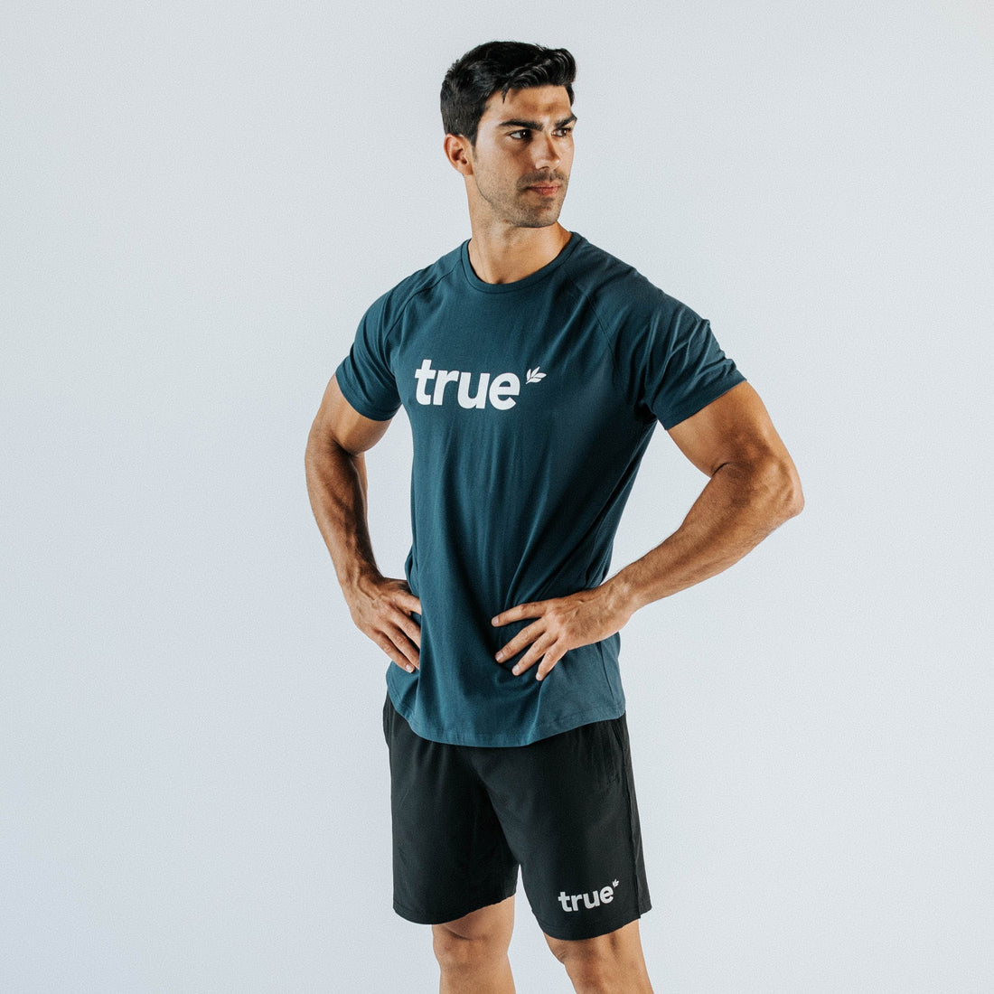 Male model wearing navy "true" logo crewneck t-shirt and black "true" athletic shorts, slim athletic fit on white studio background — men&