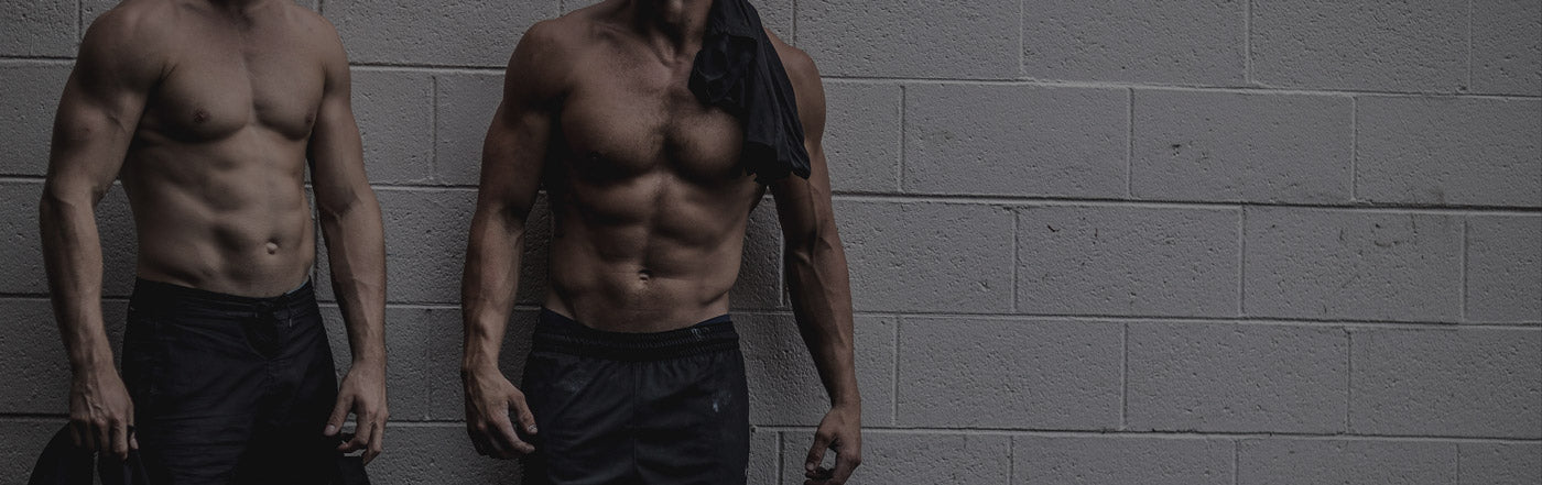 Two shirtless muscular men showing defined abs and arms standing against a gray cinderblock wall — men's fitness, weight loss and muscle-building workout concept for gym training and personal training services
