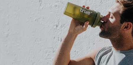 Man drinking from green True® reusable shaker water bottle against sunlit white wall — outdoor fitness hydration image for sports, gym, healthy lifestyle.