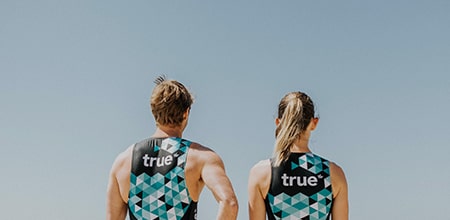 Two athletes seen from behind wearing matching teal geometric triathlon suits with "true" logo, standing under clear blue sky — fitness apparel and team kit for outdoor training.