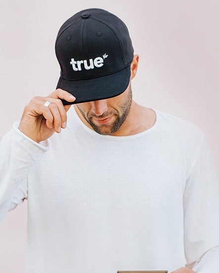 Man wearing white long-sleeve shirt and black baseball cap embroidered with "true" logo, looking down against a pale background — product shot for wholesale/online apparel banner.