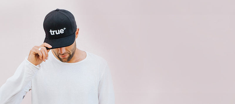 Man wearing black "true" logo baseball cap and white long-sleeve shirt against pale pink studio background, lifestyle apparel product photo for wholesale and retail display