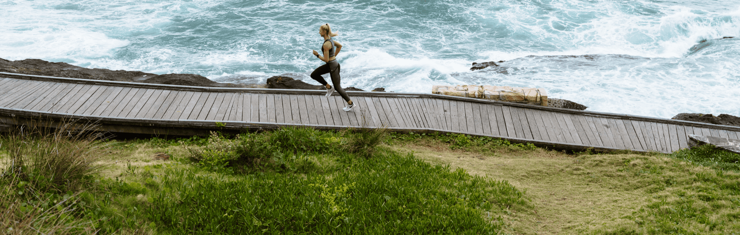 Woman runner jogging on a wooden coastal boardwalk beside rocky shore and crashing ocean waves, outdoor seaside fitness scene with green coastal grass.