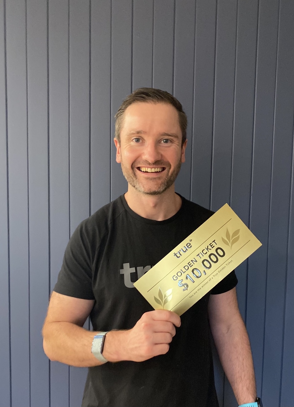 Smiling man holding a gold "True" Golden Ticket voucher for $10,000, portrait-style promotional photo against a vertical blue paneled wall.