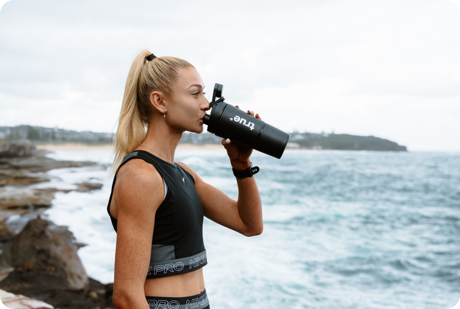Young woman in black Nike Pro workout clothes drinking from a black "True" shaker bottle on a rocky ocean coastline, outdoor fitness hydration by the sea.