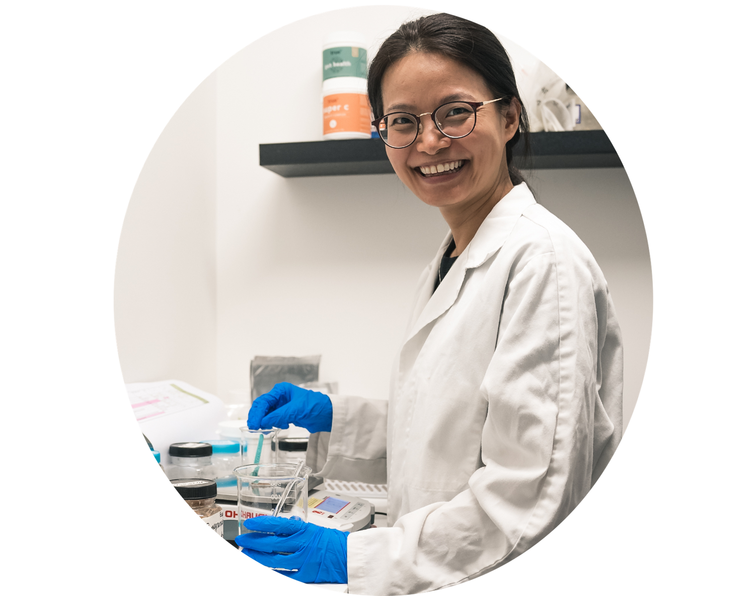Smiling female scientist in a white lab coat and blue gloves mixing a solution in a beaker at a clinical laboratory bench.