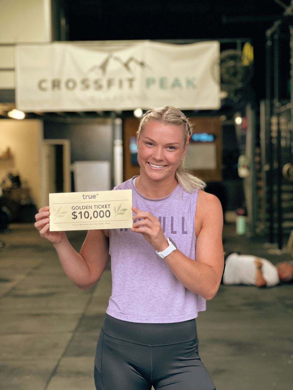 Smiling young woman in workout clothes holding a "true golden ticket $10,000" voucher inside CrossFit Peak gym, ropes and weight racks in background.