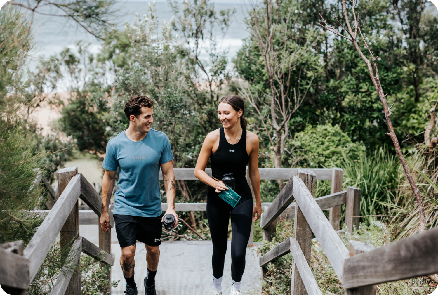 Man and woman in activewear walking up wooden beach stairs through coastal bushland, holding water bottles—outdoor fitness walk near a sandy ocean shoreline.