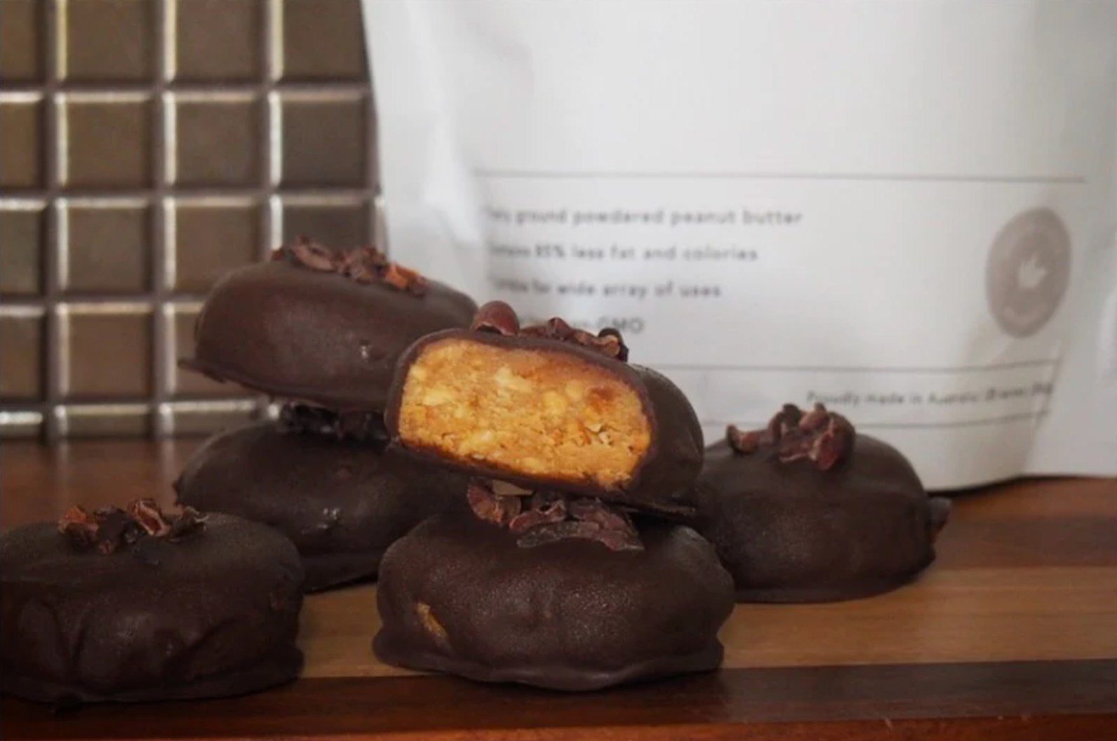 Chocolate-coated peanut butter cookies topped with cacao nibs, one cut open to show golden peanut butter center, on wooden board with packaging labeled “Proudly made in Australia.”
