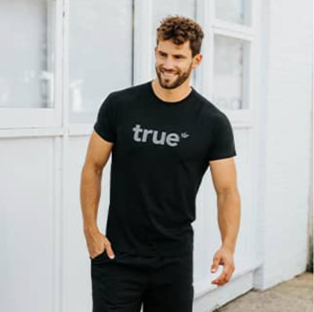 Alt text: Male model wearing a black "true" logo crewneck t-shirt and black pants, smiling and walking on an urban sidewalk in front of white building — men's casual streetwear tee product photo, responsive fit, summer outfit.