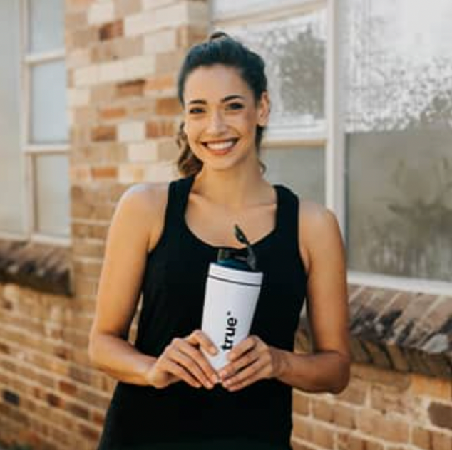 Smiling young woman in a black tank top holding a white reusable shaker bottle branded “true” while standing outdoors by a brick building window — fitness lifestyle, hydration, eco-friendly water bottle, urban wellness.
