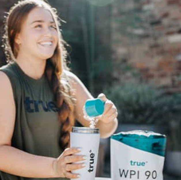 Smiling woman outdoors pouring True WPI 90 whey protein isolate powder into a white True shaker bottle on a sunny patio