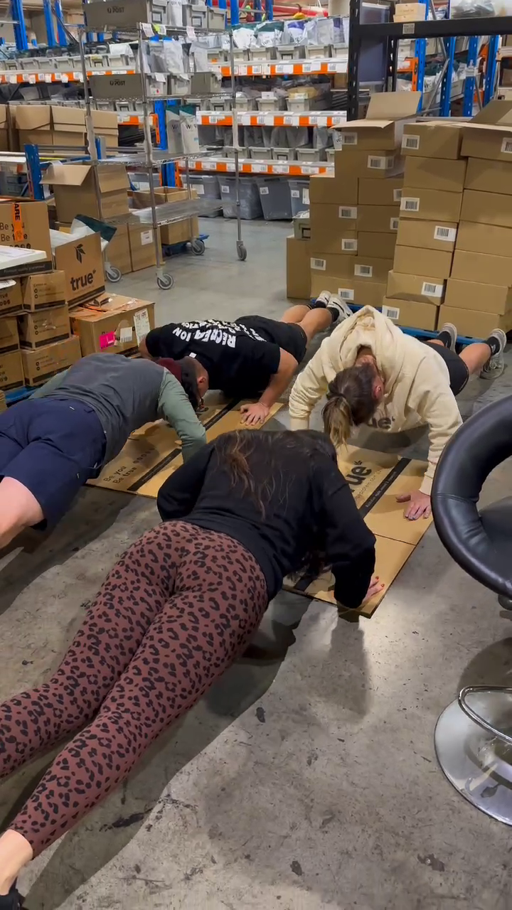 Four warehouse workers doing a push-up challenge on flattened cardboard in a busy fulfillment center aisle surrounded by stacked boxes, metal shelving and rolling carts.